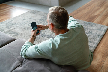 Mature man using smart phone leaning on sofa in living room at home