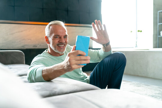 Smiling man on video call waving at smart phone in living room
