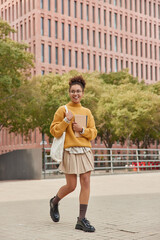 Fototapeta na wymiar Full length shot of beautiful curly haired female student wears yellow knitted jumper skirt and black shoes carries fabric bag holds notepads strolls in city centre looks away with glad expression.
