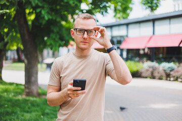 Portrait of handsome mature businessman holding messenger bag, wearing stylish eyeglasses looking at camera. Smiling middle aged man using smartphone, standing on the street. Mobile banking concept