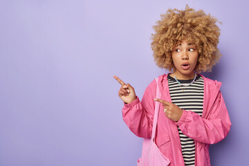 Horizontal shot of curly haired European woman dressed in striped jumper and pink jacket points away on left corner isolated over purple background demonstrates banner new promo deal or logo.