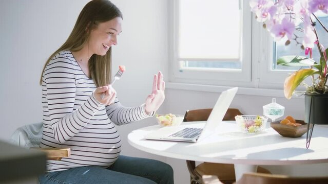 Pregnant woman sitting at table at home using laptop for video call. - Powered by Adobe