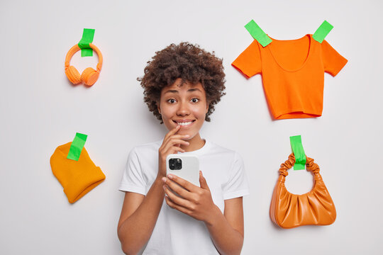 Waist up shot of positive pretty young woman holds mobile phone checks notification bites lips awaits for something poses against white background with plastered orange clothes and accessories