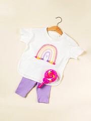 A children's T-shirt with a rainbow on a hanger and lilac shorts on a beige background. Children's clothing for summer.