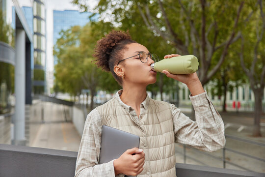 Horizontal shot of curly haired woman feels thirsty drinks water from bottle carries digital notepad wears round spectacles shirt vest poses outdoors against street background. People and lifestyle