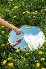 nature concept - hand touching sky reflection in round mirror on summer field