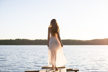 Beautiful Russian girl dressed in a white dress, walking along a wooden pier on the bank of a river...