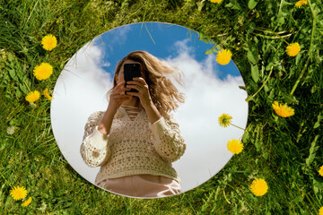 nature and people concept - woman with smartphone reflection in round mirror on summer field with dandelion flowers - Powered by Adobe