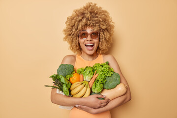 Positive beautiful woman embraces bunch of fresh green vegetables and fruits wears casual t shirt...