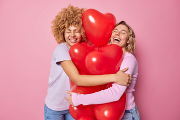 Cheerful optimistic women embrace big bunch of heart shaped inflated balloons dressed in casual...
