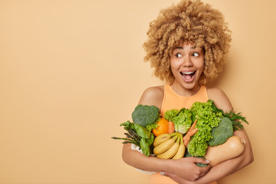 Indoor Shot Of Positive Surprised Young Woman Has Curly Bushy Hair Embraces Variety Of Fresh Green Vegetables And Fruits Returns From Supermarket Isolated Over Beige Background Empty Space For Text