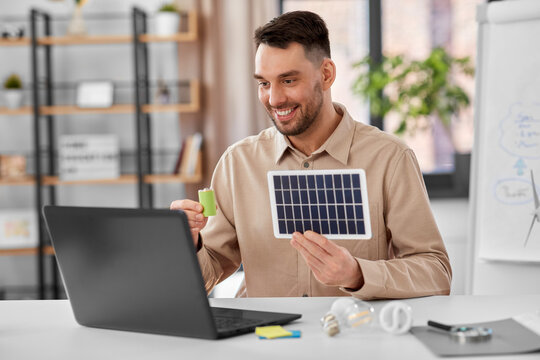 distance education, school and green energy concept - happy smiling male teacher with laptop computer and solar battery model having online class at home office