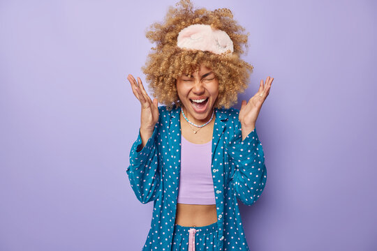 Emotional curly haired beautiful woman dressed in blue comfortable pajama sleepmask raises hands exclaims loudly wakes up early in morning isolated over purple background. People and emotions concept