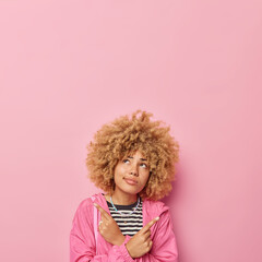 Hesitant curly haired woman points index fingers sideways dressed in jacket chooses between two options focused above thoughtfully isolated over pink background blank space for advertisement