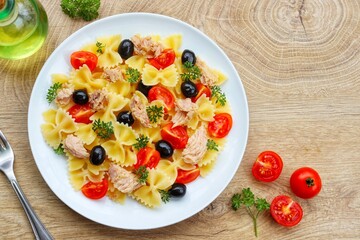Farfalle pasta salad with canned tuna in olive oil,black olives,cherry tomatoes,parsley,olive oil and peppers on plate with wooden background.Healthy Italian summer salad.Top view.Copy space