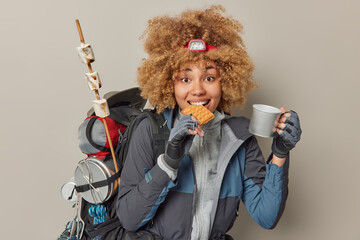 Indoor shot of pretty woman hiker has snack during journey bites waffle and drinks tea dressed in...