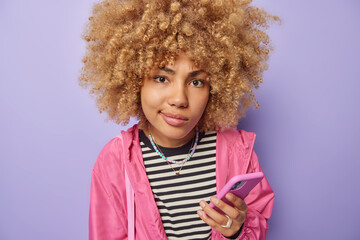 Photo of serious woman with curly hair purses lips looks attentively at camera holds mobile phone thinks about something dressed casually isolated over purple background. Cellular technology
