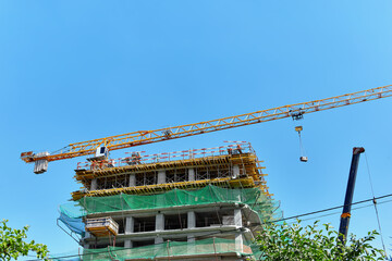 Naklejka premium A yellow tower crane lifts a cargo on a building construction site with a green mesh against a clear blue sky