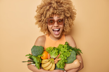 Optimistic glad young woman with curly hair carries fresh spring vegetables returns from market exclaims loudly wears sunglasses and t shirt isolated over beige background. Green grocery concept
