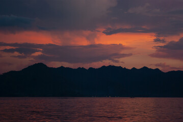 Fototapeta na wymiar Dark majestic purple sunset on ocean with dark silhouette mountains as line, orange, pink saturated reflection sunbeams in calm water. Dramatis indonesian landscape in evening, beautiful trip in asia.