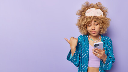 Photo of displeased curly haired European woman has worried expression checks notification via mobile phone dressed in pajama points thumb away at blank space isolated over purple background