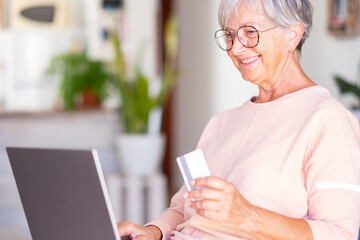 Fototapeta na wymiar Elderly woman browsing by laptop enjoying shopping online. Joyful and smiling beautiful senior lady indoors being in great mood using credit card to purchase