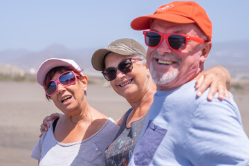 Happy group of mature caucasian people with caps and sunglasses enjoying summer holiday at the beach, hugging and looking at camera