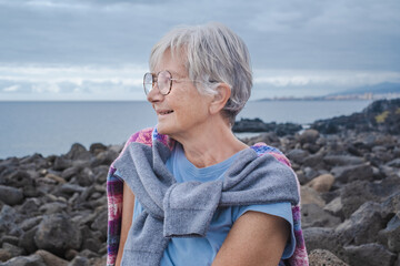 Portrait of senior gray-haired woman sitting on the cliff looking at horizon over sea. Cloudy sky in background, copy space