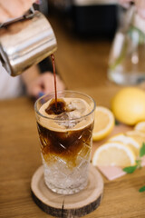 Barista pouring espresso into a glass with iced tonic, standing on the wooden bar counter with lemons on the background