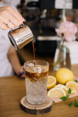 Barista pouring espresso into a glass with iced tonic, standing on the wooden bar counter with lemons on the background