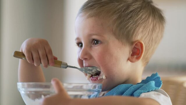 Baby eats cereal porridge. Bboy is holding spoon in his hand. Child sits at table and eats porridge.Family breakfast in kitchen. Feeding child at table.Child proper nutrition.Breakfast in kindergarten