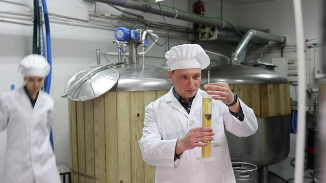 Interested brewery owner checking alcoholic content in craft beer, using test tube and hydrometer while standing near fermenters in workshop