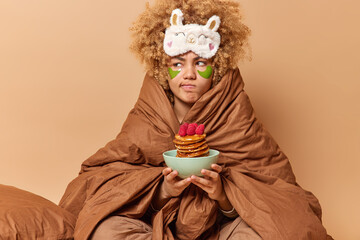 Serious discontent woman with curly hair wrapped in soft blanket holds bowl of pancakes with...