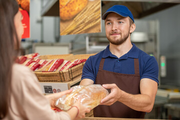 Modern grocery store worker wearing uniform helping young woman to choose tasty bread
