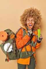 Vertical shot of positive woman with curly hair holds magnifying glass over mouth shows white teeth holds toothbrush and toothpaste dressed in windbreaker carries rucsack. Exploration and camping - obrazy, fototapety, plakaty
