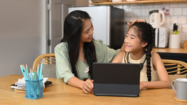Caring asian mom helping her daughter doing homework, studying online in kitchen