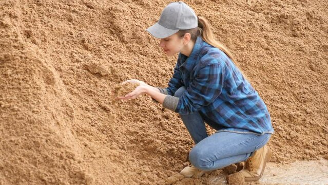 Young female farmer holding handful of beer bagasse while squatting near pile in storage area outdoors at dairy farm, checking quality of organic livestock feed. High quality 4k footage