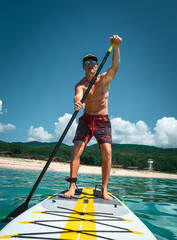 Tanned man stands on a yellow paddle board and paddles. Active sport on the beach in the turquoise sea water - obrazy, fototapety, plakaty