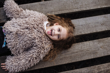 Portrait of a happy little girl showing tongue lying on a wooden background. Beautiful baby enjoys a walk in the park. Fashionable children.