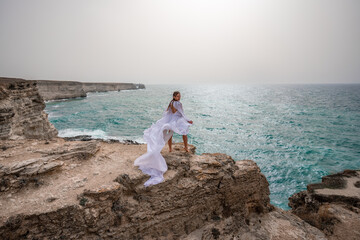 Fototapeta na wymiar Happy freedom woman on the beach enjoying and posing in white dress over the sea. View of a girl in a fluttering white dress in the wind. Holidays, holidays at sea.