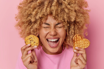 Headshot of joyful European woman with curly hair holds delicious waffle and cookie enjoys eating...