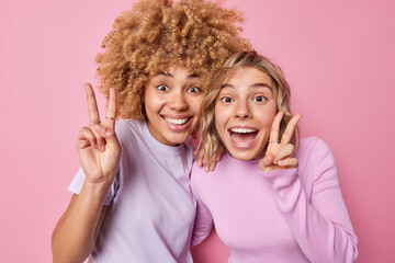 Positive happy women have fun carefree mood make peace gesture or v sign have glad expressions stand closely dressed in casual clothes isolated over pink background express optimism and joy.