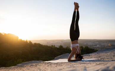 Beautiful young caucasian girl doing yoga and performing a headstand on a rock on a background of sunset and cityscape