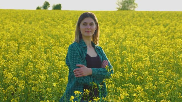 beautiful smiling farmer woman standing yellow flowering rapeseed crossed her arms and looking at camera. concept of female farmer.