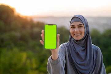 Beautiful young girl in a hijab holding a phone with a green screen on a background of the landscape at sunset