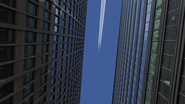 Airplane Flies over Office Skyscrapers on Blue Clear Sky Leaving White Traces