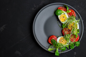 Salad of boiled eggs, herbs and tomatoes on a gray plate, top view. Place for text. Creative food on a black background.