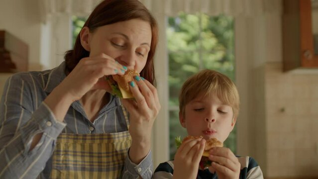 Mother and son eating homemade burgers in the kitchen. Happy family enjoying cheeseburgers together at home. Handheld camera.