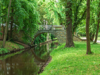 Park dworski w mieście Iłowa w Polsce. Płynąca leniwie przez park rzeka. Nad rzeką zabytkowy, kamienny mostek. Na brzegu zielona trawa, wokół drzewa.