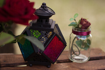 Obraz na płótnie Canvas Black lantern in stained glass. Near a transparent jar with a rose inside. They are on a wooden box. Next to vintage wall